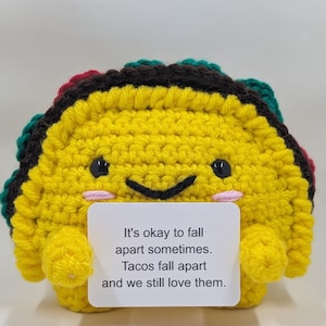 May include: A crocheted taco with a smiling face, in shades of yellow, green, brown, and red. The taco holds a small card with the text: "It's okay to fall apart sometimes. Tacos fall apart and we still love them."