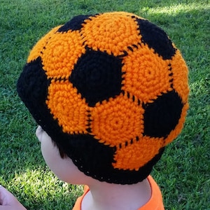 Könnte beinhalten: Eine gehäkelte Mütze in Form eines Fußballs mit schwarzen und orangefarbenen Feldern.