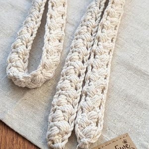 May include: Cream-colored macrame lanyard with a woven design. The lanyard has silver-colored swivel clasps and a clear plastic tag holder. A small brown tag with the text "Bee Ewe Designs" and "100% Cotton" is also visible.