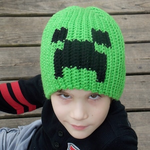 May include: A child wearing a green knitted hat with a black creeper face design. The hat is crocheted and has a ribbed texture.
