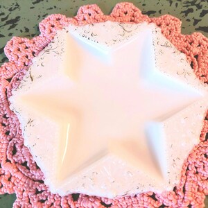 May include: A white star-shaped dish with a delicate floral pattern, set on a pink crocheted doily.
