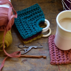 May include: A collection of handmade crochet items, including coasters and yarn. The coasters are in shades of teal, mauve, and olive green, with wooden ring handles. A white mug with a pink base sits on a coaster. Other items include scissors and a crochet hook.