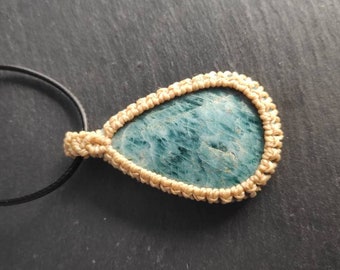 May include: Teardrop-shaped pendant necklace featuring a polished, teal-colored stone. The stone is encased in a beige macrame setting and hangs from a black cord with a silver-tone clasp. A unique, handcrafted jewelry piece.
