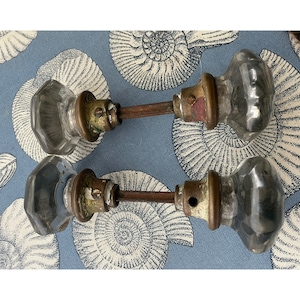 May include: Two antique glass doorknobs with brass hardware and a connecting rod. The knobs are faceted and clear, showing signs of age. The brass has a tarnished appearance. The background is a blue fabric with a white seashell pattern.