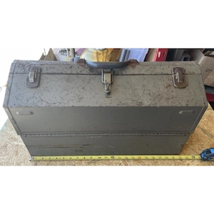 May include: A vintage, gray metal toolbox with a handle and clasps. The toolbox is rectangular, with a slightly worn appearance. A measuring tape is placed next to the toolbox, showing a length of approximately 12 inches.
