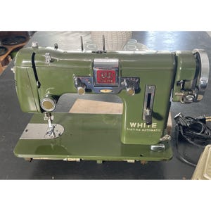May include: An olive green White brand sewing machine with chrome accents. The machine has a flat base, a needle, and a hand wheel. The text "WHITE" and "Z/G14G AUTOMATIC" are visible on the machine.