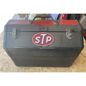 May include: A vintage metal toolbox with a red handle and trim. The toolbox is dark gray with an "STP" logo sticker on the front. It has two metal latches and a handle for carrying. The toolbox appears to be well-used.