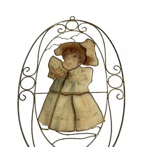 Puede incluir: Un adorno de pared ovalado antiguo con una imagen pintada de una niña con vestido y sombrero. La imagen está enmarcada por una filigrana metálica decorativa envejecida con volutas y acentos circulares. El vestido de la niña es de color crema claro.