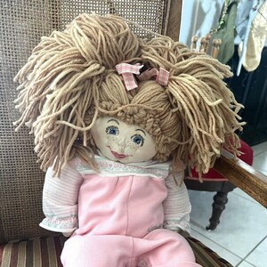 May include: A handmade rag doll with yarn hair, wearing a pink outfit with a floral print top and overalls. The doll has painted blue eyes and a smiling face.