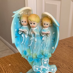 Puede incluir: Tres ángeles azules con alas, sosteniendo libros y rezando, de pie sobre una base azul con una cruz blanca y rosas azules.