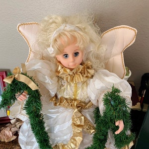 May include: A white and gold angel doll with blonde hair and blue eyes, wearing a white dress with gold trim and holding two green garlands.
