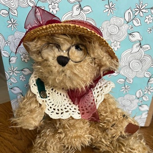 May include: A brown teddy bear wearing a straw hat with a red ribbon, round glasses, and a white crocheted collar. The bear is sitting on a wooden surface.