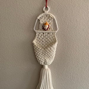 May include: White macrame wall hanging with a clown face detail and a tassel.