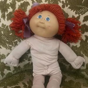 May include: A vintage doll with a soft, fabric body and a vinyl head. The doll has bright blue eyes, a small nose, and a slight smile. Her red yarn hair is styled in pigtails, each tied with a purple ribbon. The doll is wearing a light pink onesie.