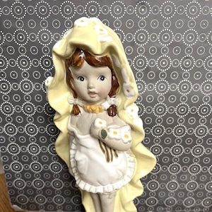 May include: A porcelain figurine of a girl wearing a yellow dress with white flowers. She is holding a bouquet of white flowers and has brown hair and blue eyes.