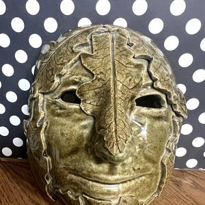Könnte beinhalten: Eine Keramikmaske mit grüner Glasur und Blattdetails. Die Maske hat einen lächelnden Ausdruck und ist so gestaltet, dass sie wie ein Gesicht aussieht.