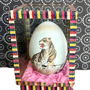 Puede incluir: Un huevo blanco pintado a mano con un tigre, exhibido en una caja de vidrio transparente con una base de terciopelo rosa. La caja tiene un borde de rayas de colores.