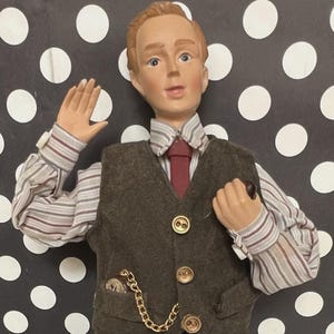 May include: A vintage doll dressed in a brown tweed suit, vest, and trousers. The doll has a striped shirt, a red tie, and black shoes. A gold chain is attached to the vest. The doll is posed with one hand raised.
