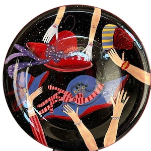 May include: A black ceramic plate featuring a hand-painted design of four hats and four arms. The hats are red, blue, and yellow with white and pink accents. The arms are painted in a whimsical style with white gloves and red and white striped sleeves.