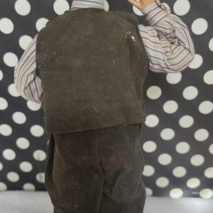 May include: A doll figurine of a man wearing a brown vest, pants, and a striped shirt. The doll has light brown hair and is posed with one arm raised. The background is black with white polka dots.