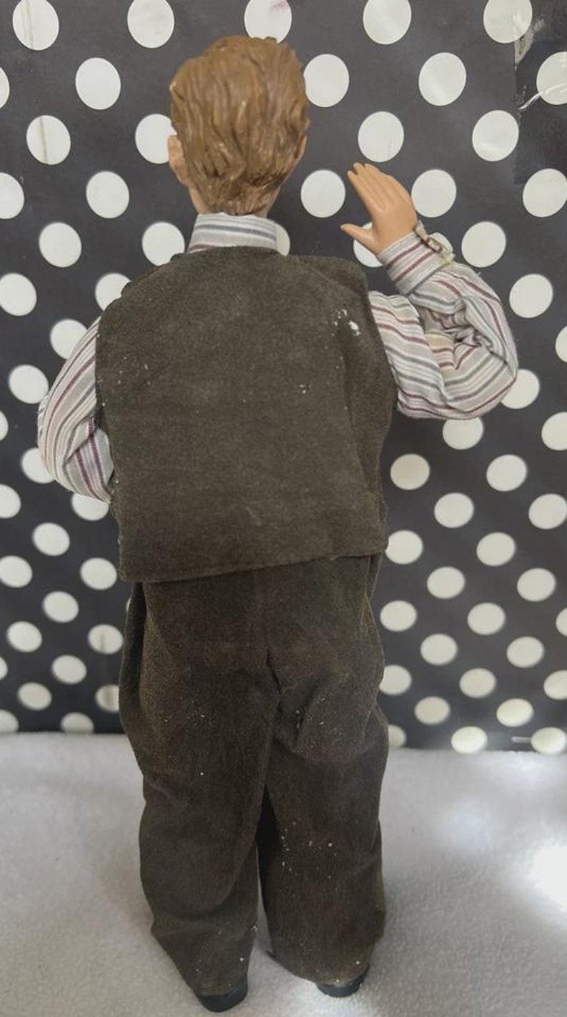 May include: A doll figurine of a man wearing a brown vest, pants, and a striped shirt. The doll has light brown hair and is posed with one arm raised. The background is black with white polka dots.