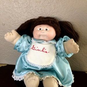May include: A Cabbage Patch Kid doll with brown hair, wearing a light blue dress with a white bib that says "Giulia".