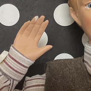 May include: A doll with a painted face, light brown hair, and blue eyes. The doll is wearing a brown jacket and a striped shirt with red and white stripes. The background features white circles on a dark gray surface.