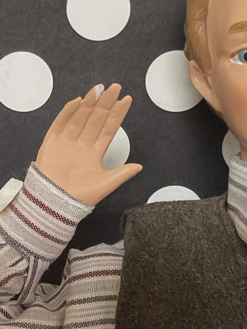 May include: A doll with a painted face, light brown hair, and blue eyes. The doll is wearing a brown jacket and a striped shirt with red and white stripes. The background features white circles on a dark gray surface.