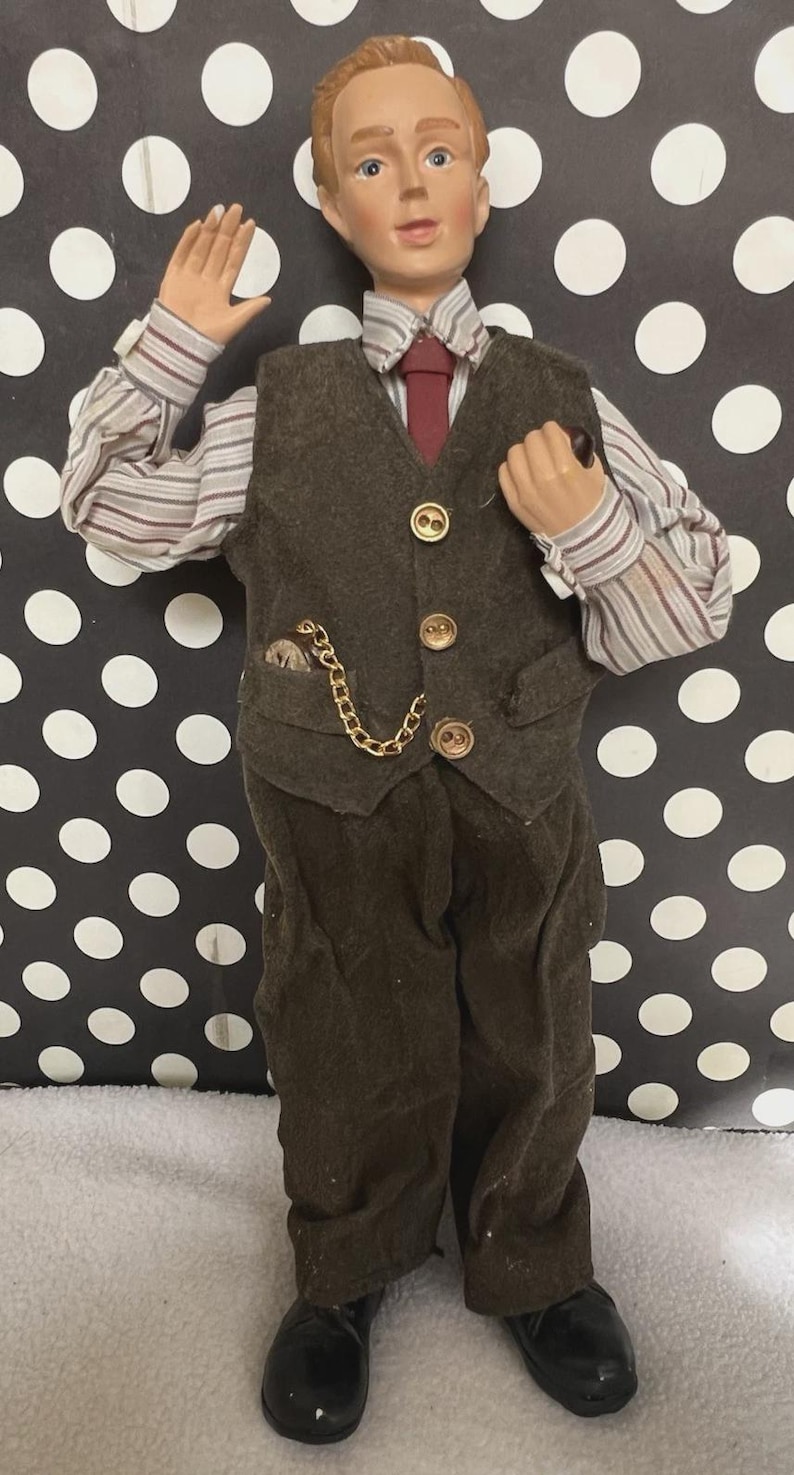 May include: A vintage doll dressed in a brown tweed suit, vest, and trousers. The doll has a striped shirt, a red tie, and black shoes. A gold chain is attached to the vest. The doll is posed with one hand raised.