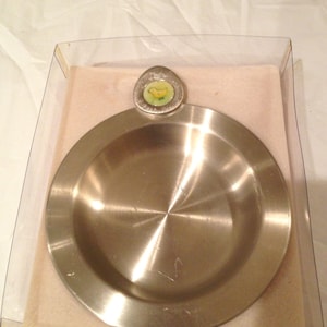 Puede incluir: Un plato de plata con un borde elevado y un pequeño diseño redondo de esmalte amarillo y verde en el centro. El plato está en una caja de plástico transparente con un fondo blanco.