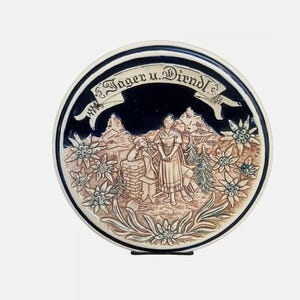 Puede incluir: Un plato redondo de cerámica con fondo azul y blanco. El plato presenta un diseño en relieve de tres figuras con vestimenta tradicional bávara de pie frente a una escena de montaña. El plato está decorado con flores de edelweiss y el texto "Jager u. Dirndl".