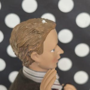 May include: A figurine of a young person with light brown hair, blue eyes, and a fair complexion. The figure wears a dark brown coat, a striped scarf, and a white shirt. The background is black with white polka dots.