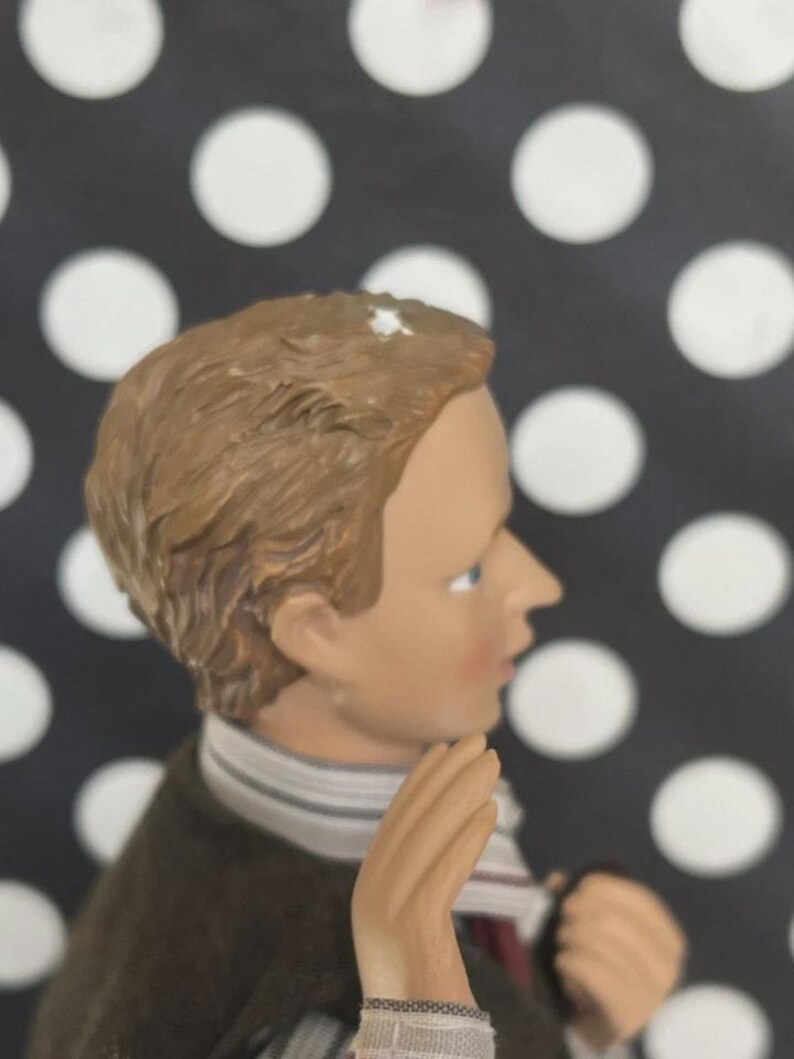 May include: A figurine of a young person with light brown hair, blue eyes, and a fair complexion. The figure wears a dark brown coat, a striped scarf, and a white shirt. The background is black with white polka dots.