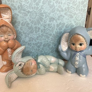 May include: Three ceramic figurines of children dressed in bunny costumes. The figurines are in various poses, including one with arms crossed, one with hands clasped, and one lying down. The figurines are painted in pastel colours, including blue, brown, and white.