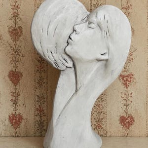 May include: A white sculpture depicting two faces in profile, positioned closely together as if in an embrace. The piece features smooth, flowing lines and a matte finish, capturing the contours of the noses, closed eyes, and hair in a unified form.