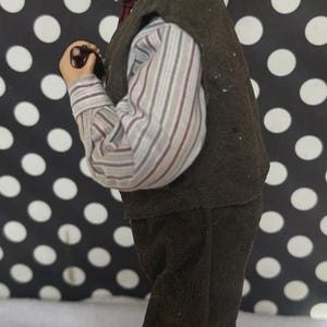 May include: A figurine of a young person in a brown vest, striped shirt, and dark pants. The figure is holding a small object and has short brown hair. The background is black with white polka dots.