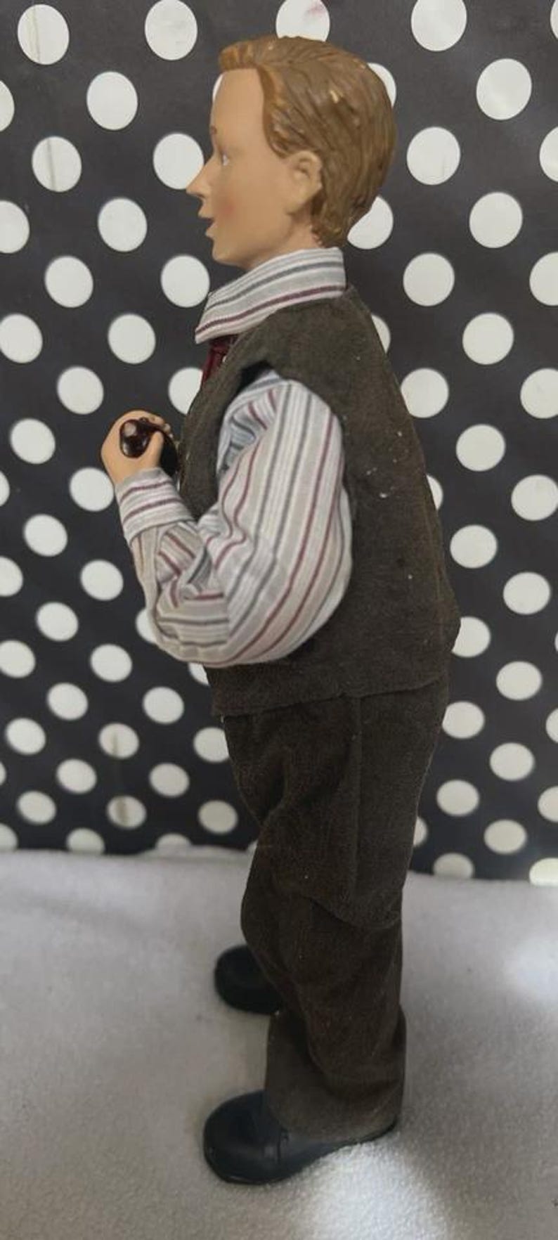 May include: A figurine of a young person in a brown vest, striped shirt, and dark pants. The figure is holding a small object and has short brown hair. The background is black with white polka dots.