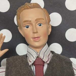 May include: A figurine of a man with fair skin, blue eyes, and light brown hair. He wears a brown vest over a striped shirt and a burgundy tie. The background is black with white polka dots.