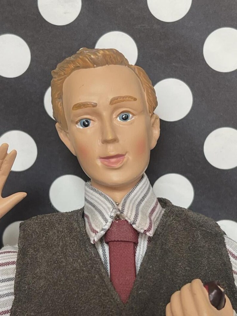 May include: A figurine of a man with fair skin, blue eyes, and light brown hair. He wears a brown vest over a striped shirt and a burgundy tie. The background is black with white polka dots.