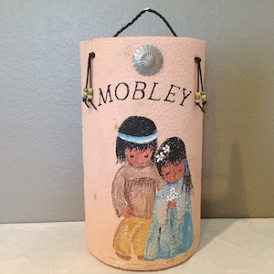 May include: A pink leather-like container with a black handle and beaded straps. The container has a painted design of two Native American children with flowers. The container is labeled "Mobley".