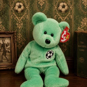 May include: A mint green plush teddy bear featuring a black stitched nose and eyes, with a small embroidered football patch on its chest. A red heart-shaped Ty tag is attached to the bear's left ear, displaying the text Ty in white.
