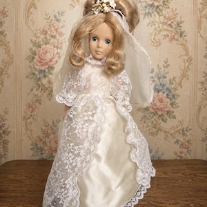 May include: A doll with blonde hair styled in an updo, wearing a white lace-trimmed dress and a matching veil adorned with small white flowers. The doll features blue eyes and a neutral expression, with the lace overlay draping over a satin skirt.