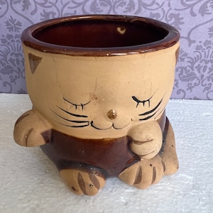 May include: A brown and tan ceramic planter shaped like a cat with closed eyes. The cat is sitting with its paws tucked under its body.