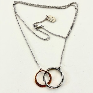 May include: A silver-toned chain necklace featuring two interlocking circular pendants. One pendant has a hammered copper-colored finish, while the other is silver with a beaded edge. A small oval tag near the clasp is engraved with AVERY 925/585.