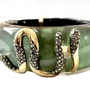 May include: A wide, translucent sage green bangle bracelet featuring a gold-toned metal snake motif that wraps around the exterior. The snake is embellished with small, dark, sparkling stones along its body and head.