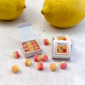 Peut inclure: Une boîte de pâtes de fruits roses et jaunes avec une étiquette qui indique "Pates de Fruits" et une image de citrons, de fraises et de framboises. La boîte est ouverte et quelques bonbons sont éparpillés autour d'elle. Il y a deux citrons en arrière-plan.