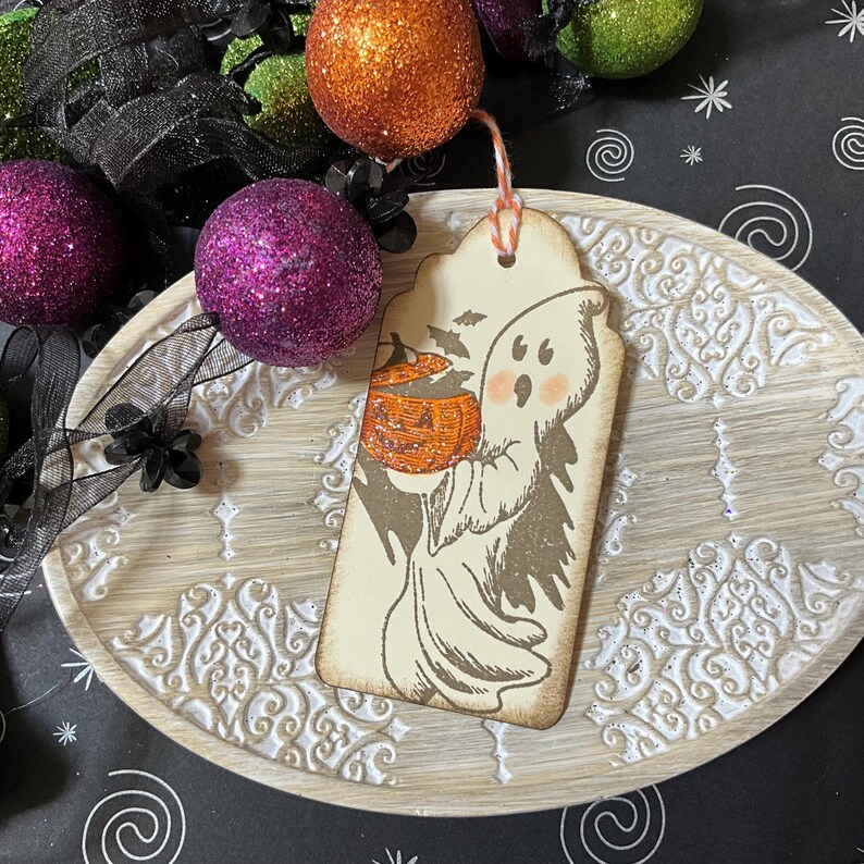 May include: A Halloween-themed gift tag featuring a cartoon ghost holding a glittery orange pumpkin. The tag has a distressed, vintage look with a string for hanging. Decorative glittery purple and orange spheres are in the background.