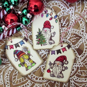 May include: Three Christmas-themed gift tags with gnome illustrations. Each tag features a different gnome design, with festive elements like a Christmas tree, string lights, and a banner. Red and white twine is attached for hanging.