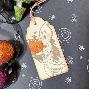 May include: A Halloween-themed gift tag featuring a friendly ghost holding a pumpkin basket. The tag has a distressed, vintage look with a beige background and a decorative orange and white string. The tag is decorated with a cute ghost illustration.