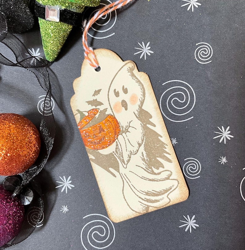 May include: A Halloween-themed gift tag featuring a friendly ghost holding a pumpkin basket. The tag has a distressed, vintage look with a beige background and a decorative orange and white string. The tag is decorated with a cute ghost illustration.
